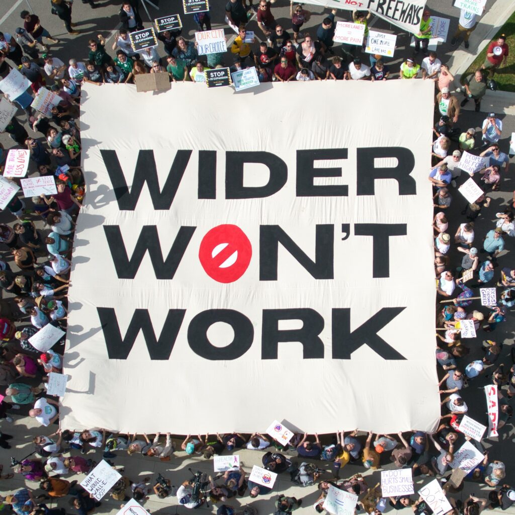 Overhead view of a crowd of advocates holding a huge sheet on which is printed boldly, wider won't work.